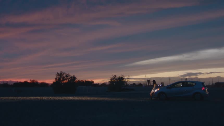A small car sits parked under a dramatic colorful sunset in a sparse desert landscape