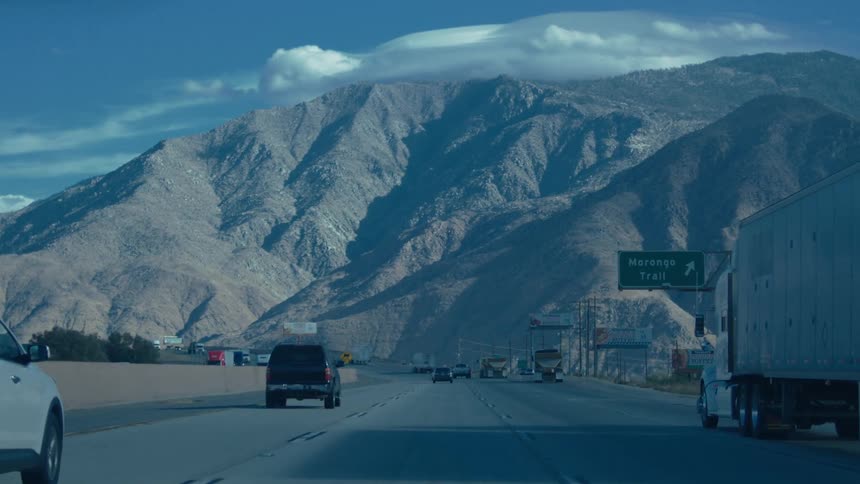 A very short video clip shows a highway with cars and trucks driving on it with large mountains in the background the sky is blue with some clouds a green road sign is visible on the right side of the road pointing to morongo trail