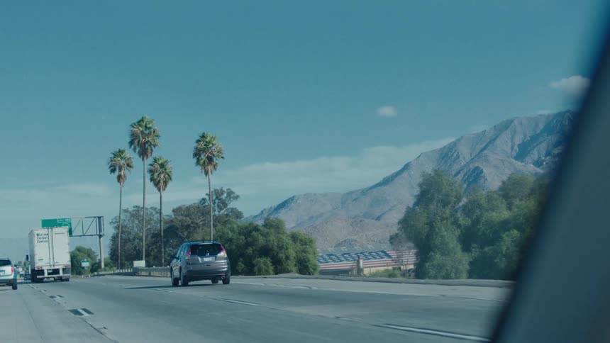 Cars cruise down a highway bordered by palm trees and mountains under a clear blue sky with a road sign visible ahead