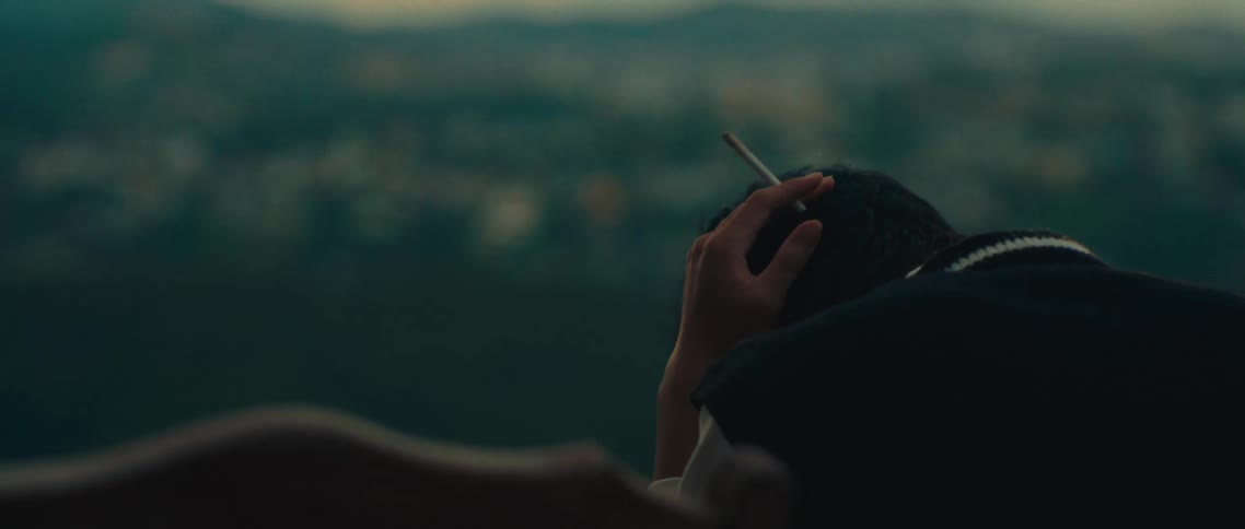A young man sitting outdoors on the outskirts of a city smoking a cigarette at midday