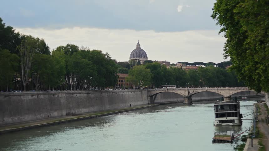 The video shows a wide shot of a river flowing through a city with a prominent domed cathedral visible in the distance a bridge crosses the river and on the right side a boat is docked at the riverbank the sky is overcast and cloudy