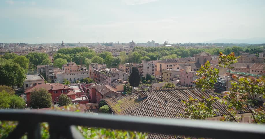 A panoramic view from behind a black metal railing slowly pans across a sprawling city with red tile roofed buildings and many green trees under a hazy sky