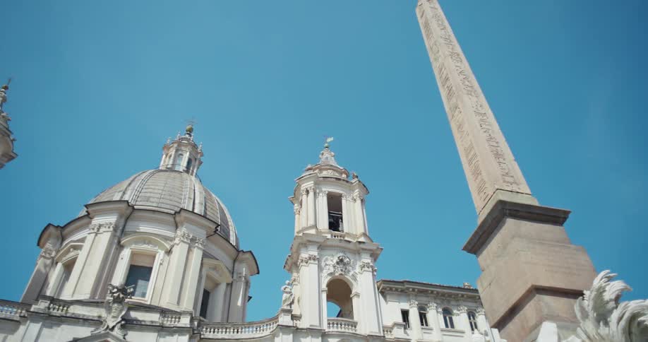 The video shows a lowangle shot of a large obelisk and a domed cathedral against a clear blue sky the obelisk is tall and slender with carvings on its side and the cathedral has a classic ornate design with a large dome and a bell tower the scene is