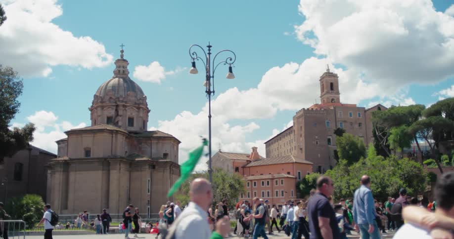 The video shows a bustling plaza with a crowd of people walking around in the background there are several large historic buildings including a prominent domed church on the left and a multistory stone building on the right the day is bright and sun
