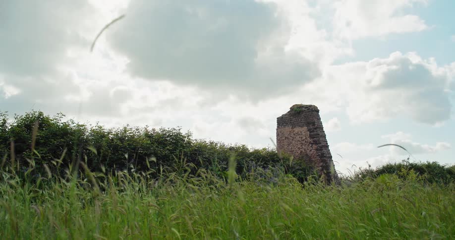 The video shows a grassy field with a large ruined brick structure in the background the structure appears to be a stone tower possibly a part of an old building or wall partially hidden behind a line of lush green bushes the sky is cloudy with a bri