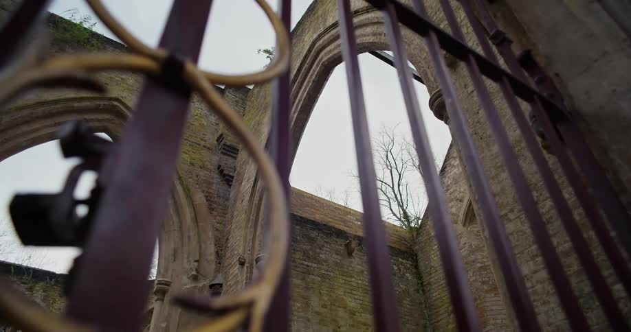 The video shows a lowangle shot looking through a set of wrought iron gates at a ruined brick building the building appears to be an old church or abbey with gothic arches and windows open to the gray sky the scene has a somber and historic feel