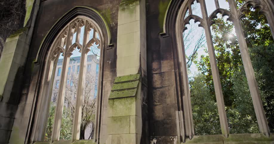 The video shows the interior of a ruined building with the camera looking through two large empty arched windows the windows are a gothic style suggesting the building was once a cathedral or monastery outside the windows trees and modern buildings
