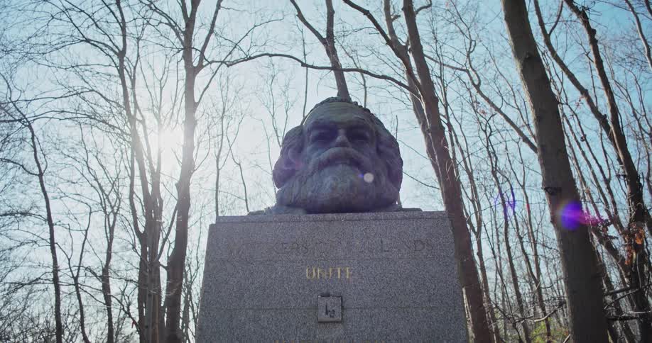 The video shows a stone bust of a man with a beard on a pedestal in a wooded area the bust is partially illuminated by sunlight filtering through the trees in the background the pedestal has the word unite carved into it