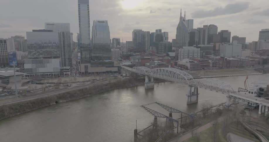 A modern city skyline rises behind a bridge spanning a wide river under a hazy sky