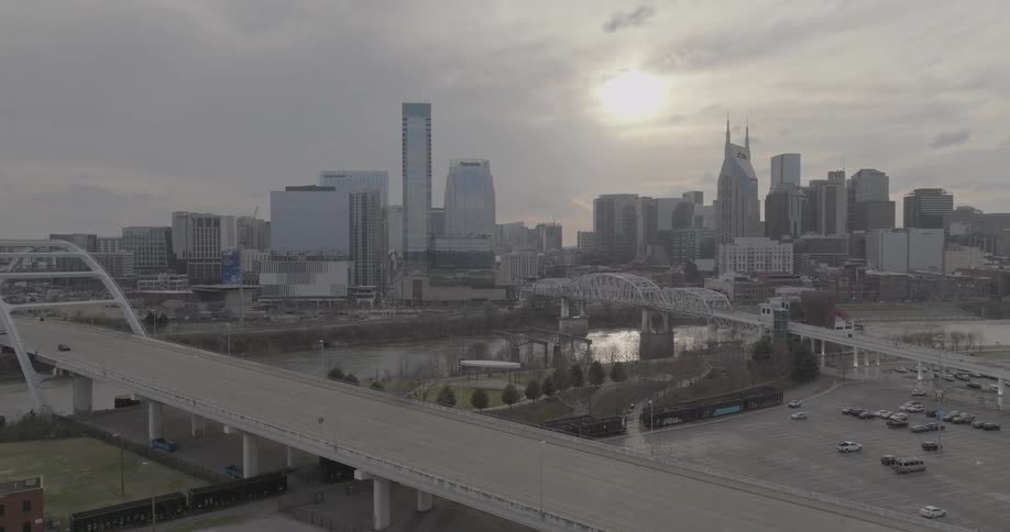 A modern arch bridge and several other bridges span a large river that winds through a city skyline under a cloudy sky with a bright sun