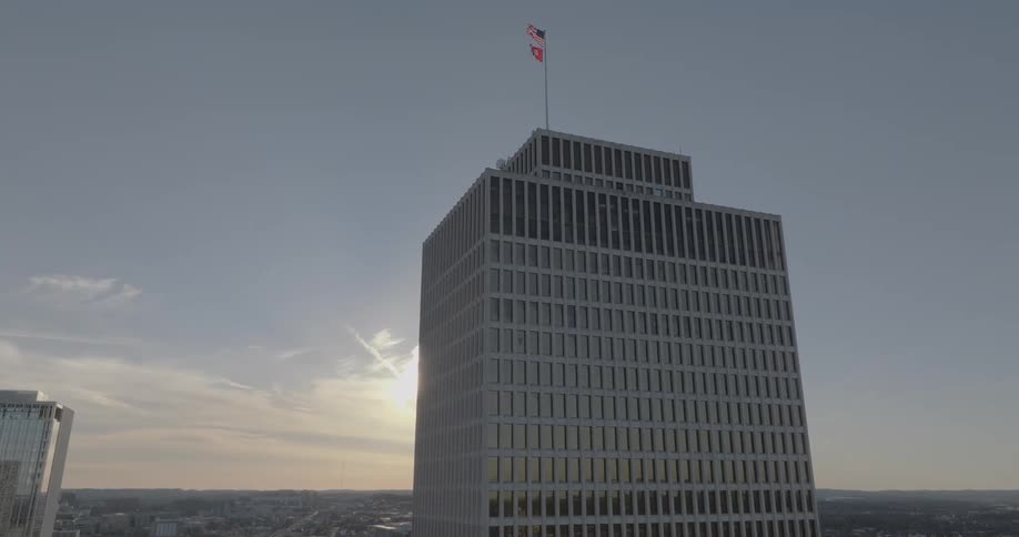 The sun sets behind a tall gray office building with many windows and two flags flying from its top with a city skyline visible in the distance