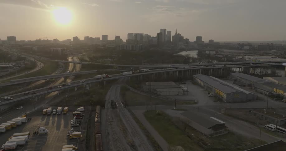 An aerial view at sunset of a busy urban transportation hub featuring a city skyline in the distance a network of highways with moving traffic and active railroad tracks with a train passing through an industrial area