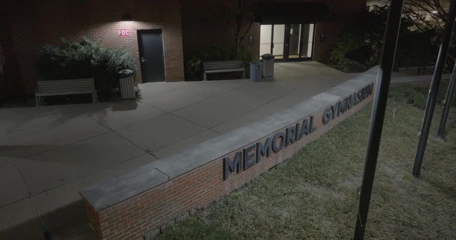 Outdoor lights illuminate a building and a half wall containing letters spelling out memorial gymnasium