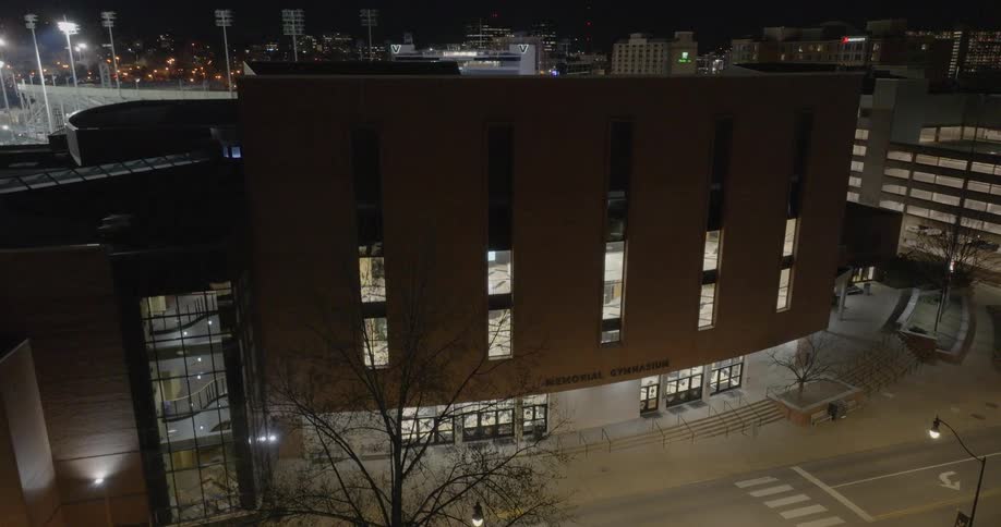 A tall brown brick building with a curved facade stands in a city at night with a university shuttle driving by on the street below