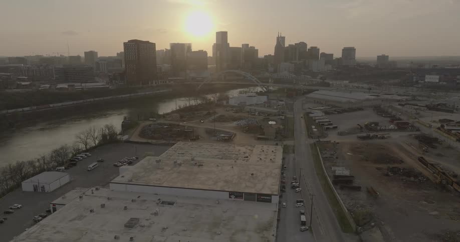 The sun sets behind a city skyline with many tall buildings which rises above a river a bridge and an industrial area in the foreground