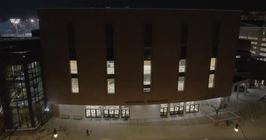 A nighttime view shows cars passing in front of memorial gymnasium its entrance warmly lit amidst the surrounding campus buildings