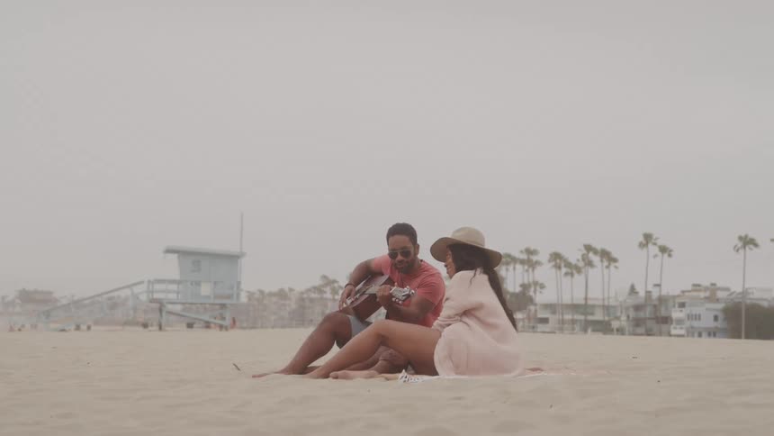 A man and a woman sitting on the beach sand with the man playing the guitar and singing to the woman