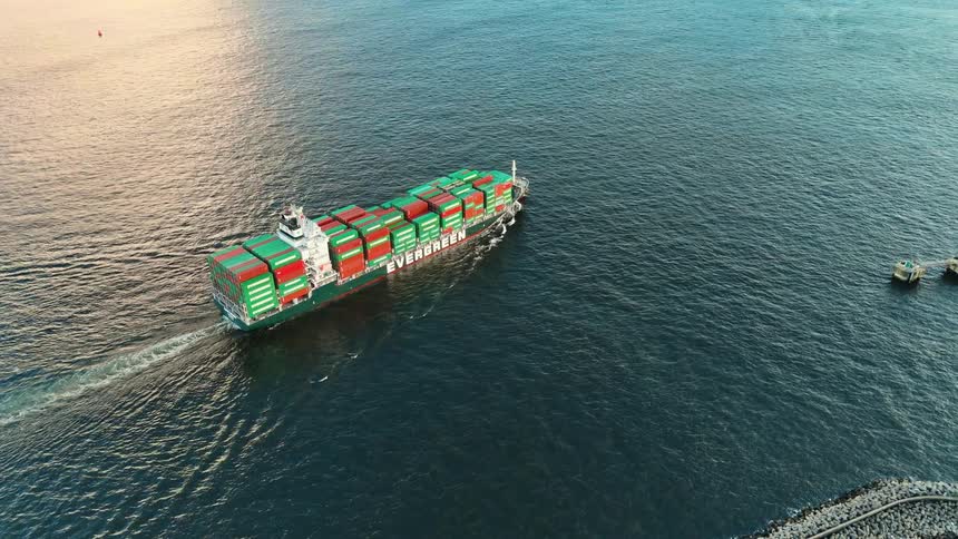 A large container ship with the name evergreen on its side moves through a dark blue body of water leaving a wake behind it with a rocky pier nearby