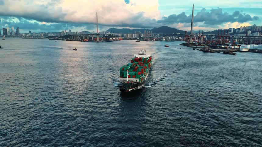 A large container ship stacked with red and green containers comes toward the camera on deep blue water with a suspension bridge and busy port in the distance