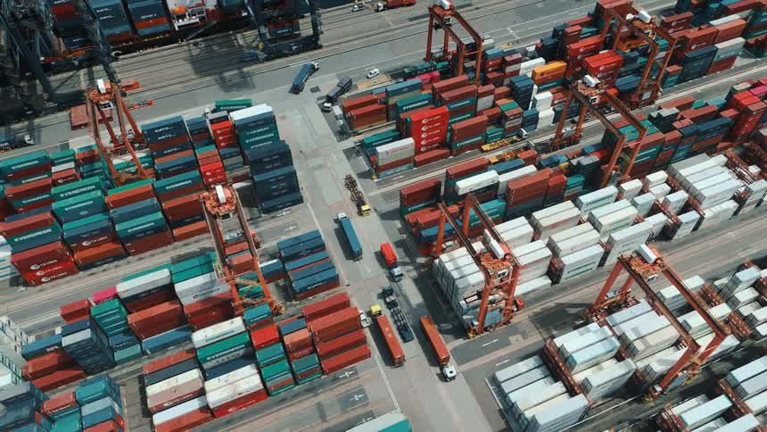 An aerial view of a busy container terminal shows stacks of multicolored shipping containers and cranes actively loading and unloading cargo beside the docked vessels