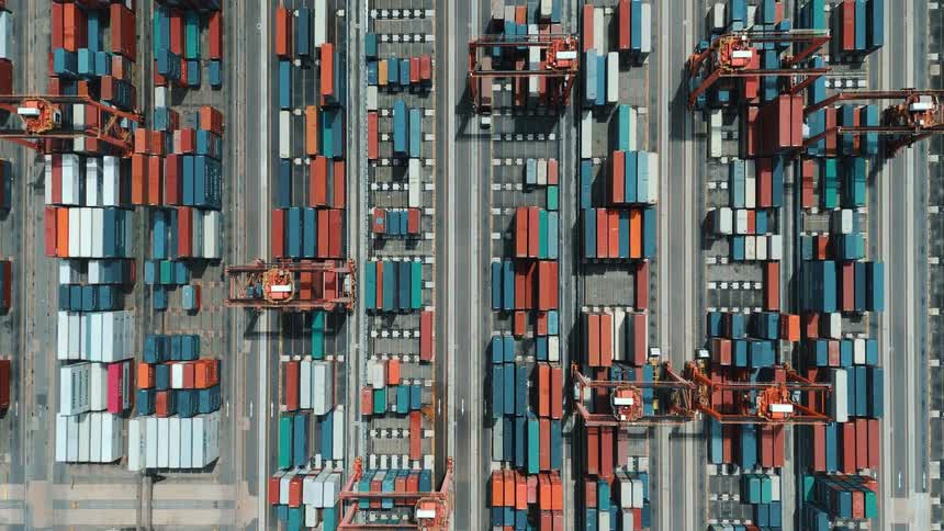 An aerial daytime view shows rows of colorful shipping containers and cranes arranged in a tightly packed port terminal