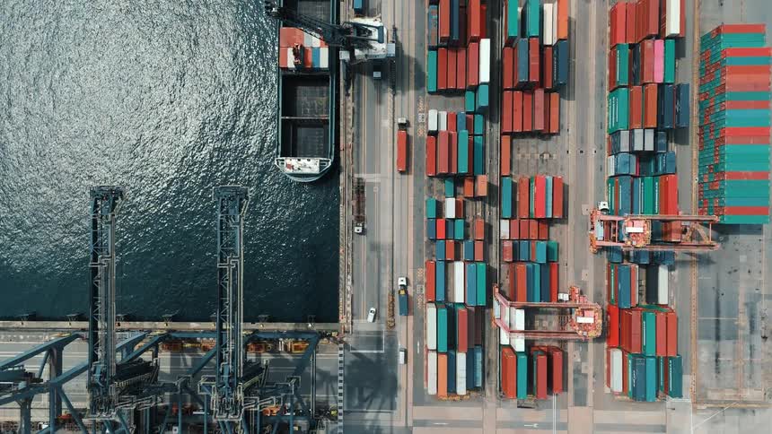 An aerial view of a busy shipping port shows rows of colorful cargo containers towering cranes and a docked ship beside shimmering water