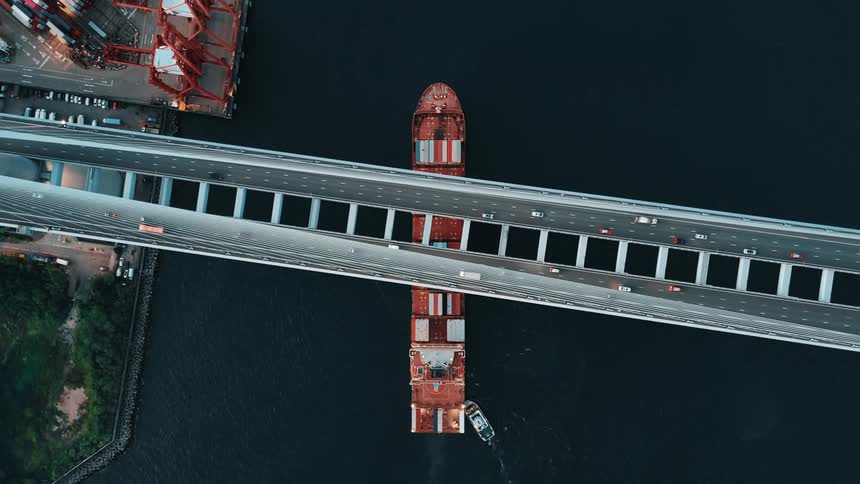 A large rusty cargo ship with a smaller white tugboat beside it passes directly under a wide highway bridge with cars on it next to a busy port