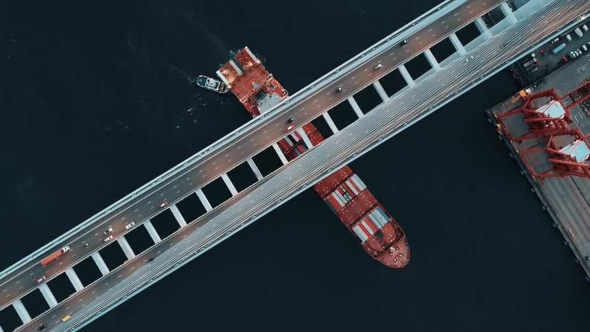 Cargo ship passing under a bridge viewed from above with vehicles moving along the roadway