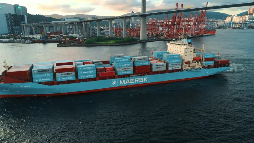 A large maersk cargo ship sails past a busy industrial port under a towering bridge