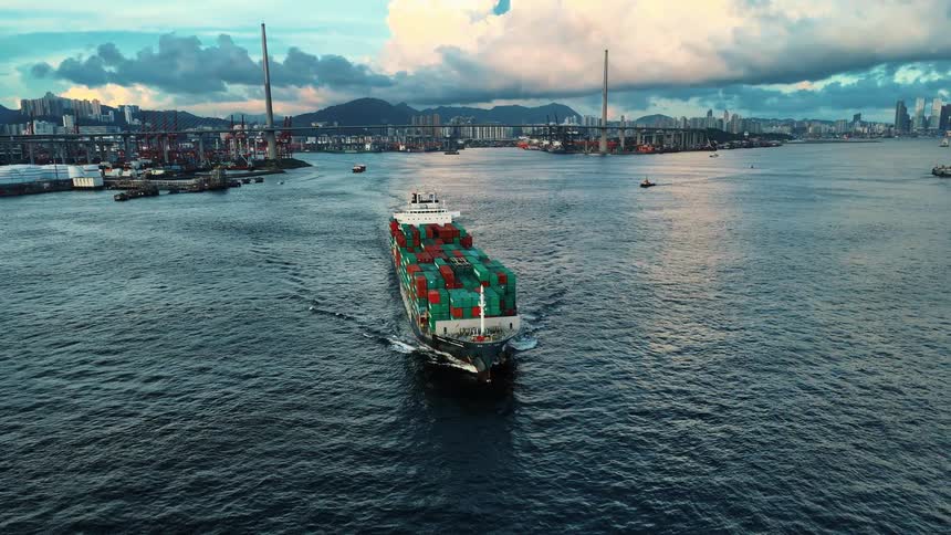 Cargo ship carrying colorful containers heading toward the port with the city and mountains in the background