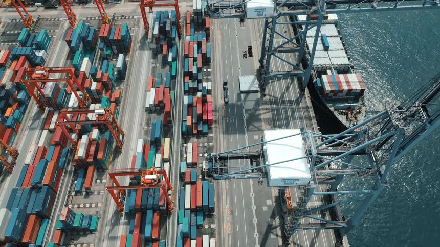 An aerial view shows rows of colorful shipping containers and large cranes at a port with a cargo ship docked alongside a wide road and deep blue water