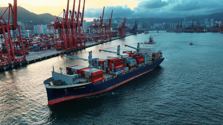 An aerial shot captures a large container ship sailing through a harbor passing a massive port terminal with numerous red cranes against a backdrop of a city skyline and distant hills at sunset