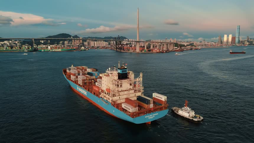 A maersk cargo ship sails through hong kong harbor at sunset guided by a tugboat with the skyline and stonecutters bridge visible in the background