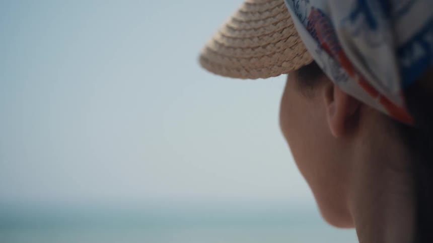 A woman in a straw hat and headscarf smiling serenely while looking out at the ocean
