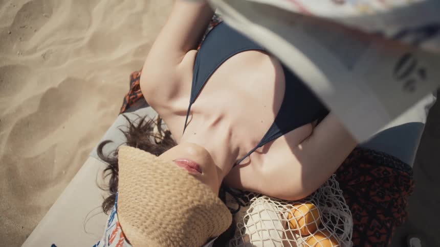 A woman wearing a dark blue swimsuit and a straw visor lies on her back on a beach chair and reads a book with a mesh bag of oranges beside her on the sand