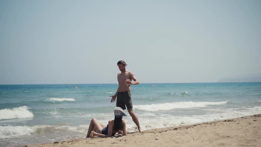 Man standing on the sand handing a drink to a woman lying by the sea