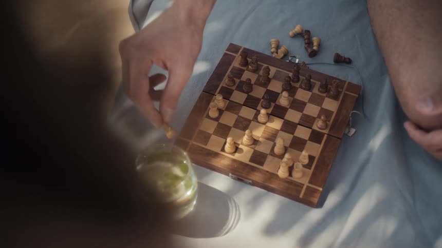 A persons hand moves a white chess piece on a small wooden board that sits on a blue blanket with other pieces scattered around and a glass with a lime slice in the foreground