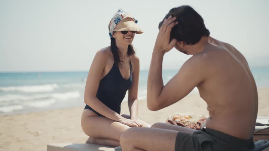 Woman in a swimsuit and straw hat smiling while talking to a man sitting on the beach