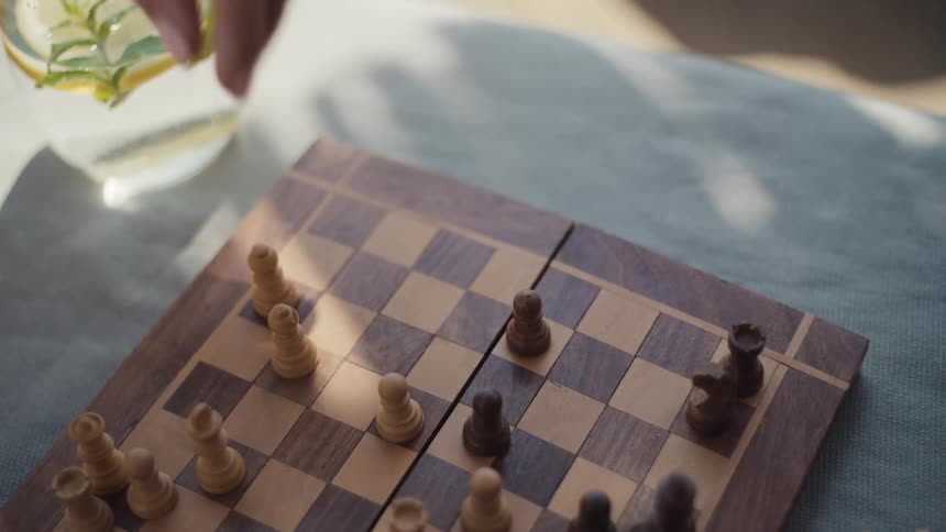 Two people sitting outdoors playing a game of chess with a glass of water on the side of the table at midday