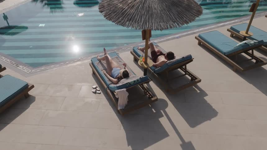 Two people lounge by a pool under a sunshade