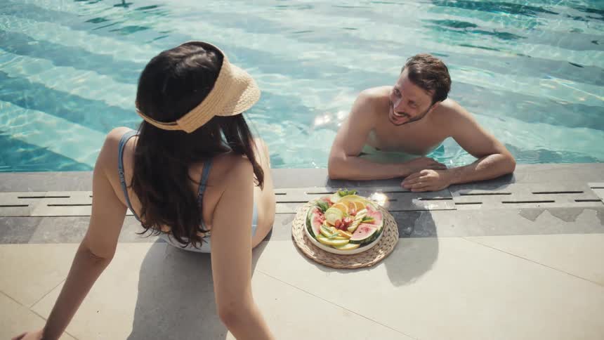 Woman sitting by the poolside with a plate of fruit beside her while talking to a man in the water