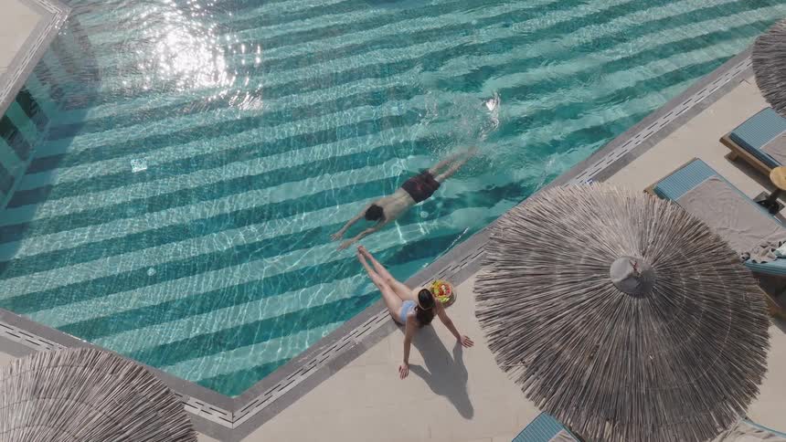 Man swims in a pool while woman sitting on the edge
