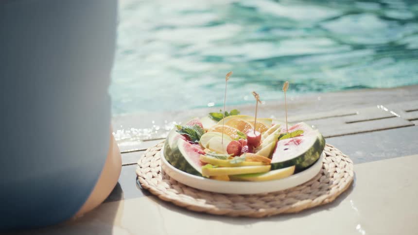 A colorful plate of fresh fruit with skewers sits poolside on a woven mat next to a person relaxing by the water