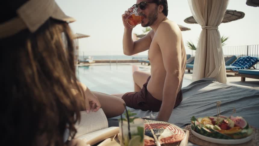 Man wearing sunglasses drinking a cocktail while lounging on a sunbed next to a woman with a book and plates of fruit