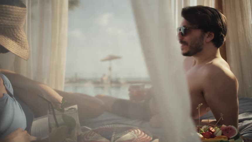 A couple is lounging in a private cabana by a pool the man is holding a drink and smiling while looking at the woman beside him