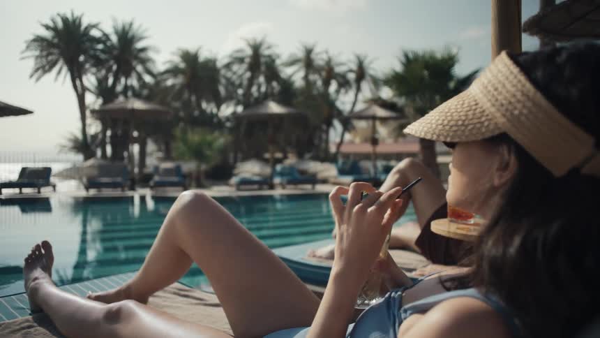 A man and woman are lounging on deck chairs under a straw umbrella by a pool in the background there is a beach with palm trees and a view of the sea
