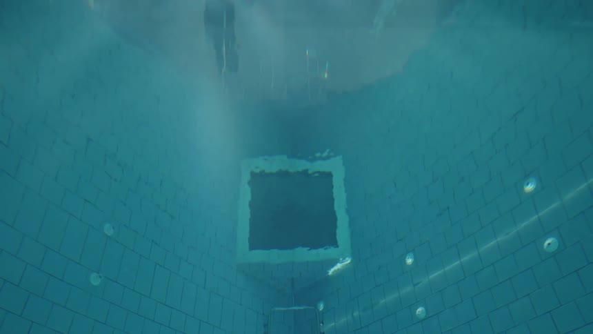 An underwater shot shows the deep end of a swimming pool with blue tiles with the main focus on a dark square opening in the pool floor