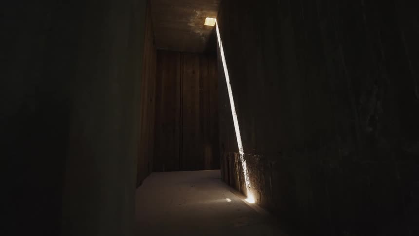 A narrow dark hallway with wooden walls is lit by a thin bright line of light that shines down one wall and onto the floor
