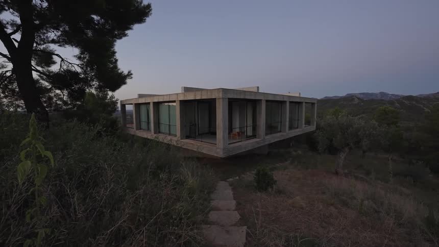 A modern geometric concrete building with large windows sits on a grassy hill with a stone path leading to its entrance under a dusky sky