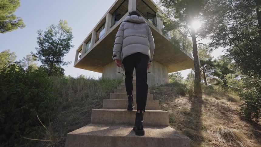 A person wearing a puffy grey jacket black pants and black boots walks up a set of concrete stairs toward a modern house with large windows